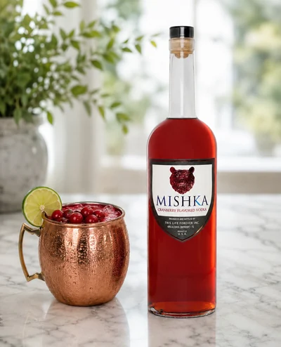 A Cranberry Crush Mule in a copper mug with fresh cranberries and a lime wheel, beside a bottle of Mishka Cranberry.